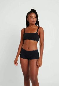 Vrouw die staat, gekleed in een zwarte crop top en bijpassende shorts, iets opzij kijkend tegen een effen lichte achtergrond.