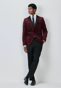 Burgunder Samtblazer mit einem strukturierten Muster, schwarzen Revers, kombiniert mit schwarzen Hosen mit rotem Seitenstreifen und polierten Schuhen.