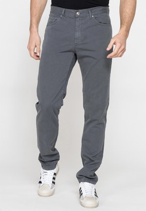 Uomo con pantaloni casual grigi e sneaker bianche con strisce nere, in piedi davanti a uno sfondo chiaro e uniforme.