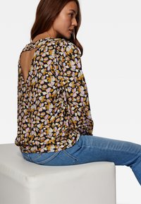 Schwarzblumiges Oberteil mit lila, orangefarbenen und gelben Mustern, verfügt über einen Rückenausschnitt und Puffärmel. Getragen mit blauen Jeans.