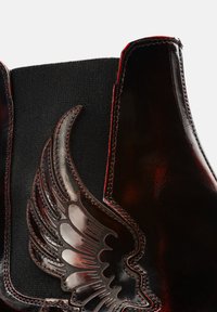 Black and dark red leather ankle boot featuring a wing design, elastic side panels, and contrasting red stitching. Smooth texture.