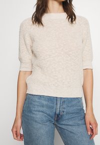 Mujer vistiendo un suéter de punto de manga corta en beige claro, combinado con unos vaqueros azul de talle alto, de pie contra un fondo liso.