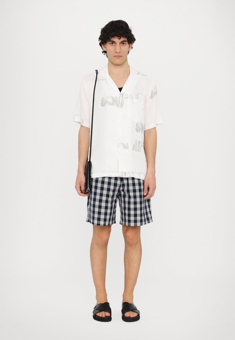 White short-sleeve shirt with grey dollar sign print, navy check patterned shorts, and black sandals, combined with a small black crossbody bag.