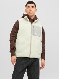 Chaleco de forro polar color beige claro con bolsillos laterales, bolsillo en el pecho con cremallera gris y un cuello alto. Lleva puesto un sudadera con capucha marrón y jeans oscuros.