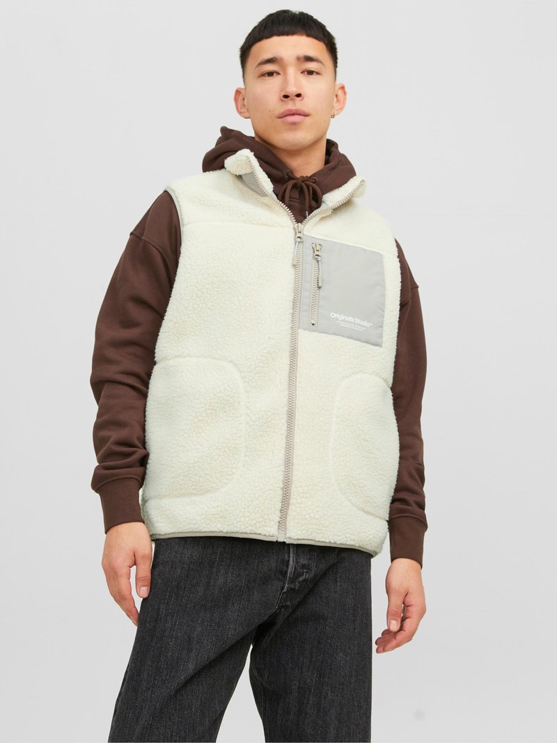 Chaleco de forro polar color beige claro con bolsillos laterales, bolsillo en el pecho con cremallera gris y un cuello alto. Lleva puesto un sudadera con capucha marrón y jeans oscuros.