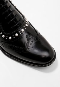 Chaussure en cuir noir avec un bout pointu, ornée de clous en argent et de perles, présentant des détails de brogue intriqués et des lacets noirs.