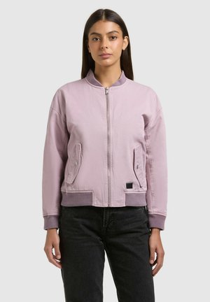 Fiatal nő egyenes sötét hajjal, világos lila cipzáras bomberdzsekit és fekete nadrágot viselve, egyszínű háttér előtt állva.