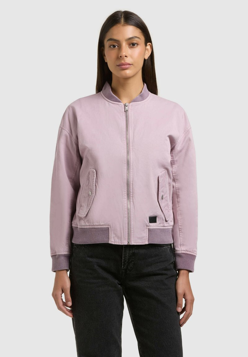 Jeune femme aux cheveux raides et foncés, portant une veste bomber zippée violet clair et un pantalon noir, debout devant un fond uni.