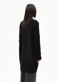 Schwarzer langer Cardigan aus weichem Strick, mit lässiger Passform, gerippten Bündchen und abgerundetem Saum, kombiniert mit grauen Hosen.
