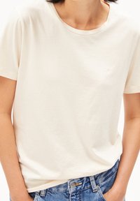 Helles beige Baumwoll-T-Shirt mit rundem Halsausschnitt, kurzen Ärmeln und einem dezenten bestickten Logo auf der Brust. Getragen mit blauen Denim-Jeans.