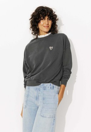 Sweatshirt gris foncé avec un col ras du cou et des poignets côtelés, présentant un cœur en imprimé léopard brodé sur la poitrine. Assorti avec un jean bleu clair.