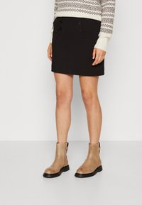 Gonna nera con dettagli a doppio bottone, abbinata a stivali alla caviglia color nocciola con zip laterale e suola nera. Maglione con motivo geometrico.