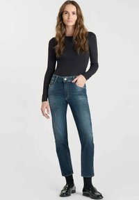 Mujer de pie vestida con una blusa negra ajustada de manga larga, jeans azules, calcetines negros y mocasines negros, contra un fondo blanco liso.