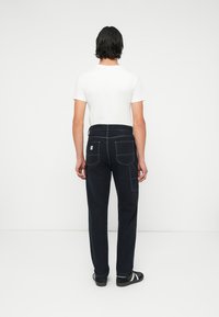Pantaloni in denim blu scuro con cuciture bianche, dotati di due tasche posteriori e di un piccolo etichetta del marchio sulla vita. Indossati con una maglietta bianca.