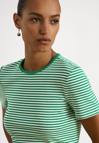 Žena so stredným odtieňom pokožky a vlasmi stiahnutými dozadu, ktorá nosí zlaté okrúhle náušnice a zeleno-bielo pruhované tričko s krátkym rukávom.