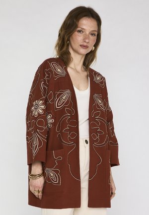 Donna con una giacca color ruggine ricamata con fiori crema, top bianco, pantaloni crema e braccialetti dorati su sfondo neutro.