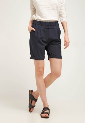 Shorts bleu marine à coupe décontractée, avec une taille élastique et des ourlets roulés, associés à des sandales noires à brides croisées.