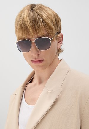 Junge Person mit hellbraunen Haaren, die große dunkle Sonnenbrillen und einen beigen Blazer über einem weißen Oberteil trägt, blickt nach vorne.