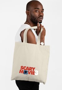 Beżowa torba na ramię z mocnymi uchwytami, z napisem "SCARY MOVIE 3" oraz grafiką w niebieskim i czarnym kolorze, trzymana przez osobę ubraną w top bez rękawów.