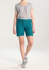 Turkosa shorts med medelhög midja, fyra fickor och knappstängning fram, kombinerat med en grå V-ringad t-shirt och grå sneakers.