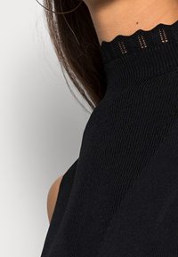 Haut noir en maille à col montant présentant une texture côtelée et un bord festonné avec un détail découpé le long du col, mettant en valeur une silhouette ajustée.