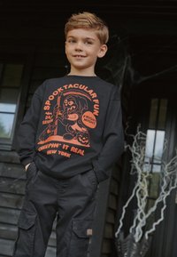 Svart långärmad T-shirt med en orange grafik som föreställer en karaktär, pumpor och text. Kombinerad med svarta cargobyxor.