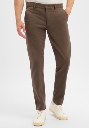 Man wearing brown slim-fit trousers and white sneakers standing against a plain white background.