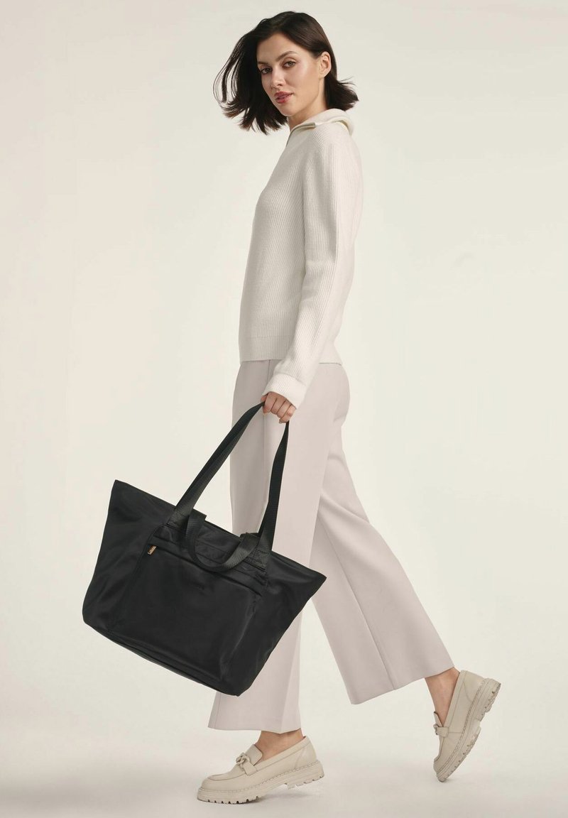 Czarna torba typu tote z podwójnymi uchwytami, wykonana z gładkiego materiału. Posiada zamek błyskawiczny na górze oraz minimalistyczny design, trzymana przez osobę w jasnym stroju.