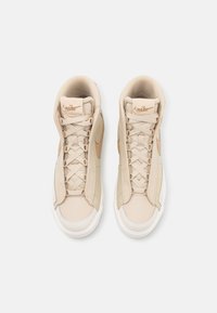 Beiga högskurna sneakers med övre del i slät läder, vadderad krage, platta snören och vit gummisula. Nike-logotyp i ljusbrunt.