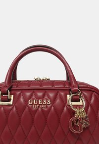 Bolso de mano de imitación cuero acolchado rojo con asas dobles, herrajes en dorado y un charm que presenta las letras "G" y "S". Logotipo de la marca: Guess EST 1981.