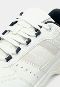 Baltas sporta apavs ar teksturētu gumijas zolīti, acs paneļiem un kontrastējošām tumšām šņórēm. Aprīkots ar polsterētu mēlīti un šuvumu detaļām.