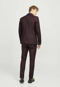 Bordeauxfarbener Anzug mit schmal geschnittenen Hosen und einem passenden Blazer, der einen Reverskragen und schwarze Knöpfe aufweist, kombiniert mit schwarzen Schuhen.