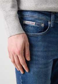 Traperice u izblijedjeloj plavoj boji, s metalnim kopčama i bijelom etiketom s natpisom "Calvin Klein." Vidljiv je rukav sivo strukturirane džemperice.