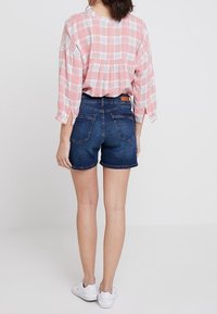 Punainen ja valkoinen ruudullinen paita, jossa on rypytetty selkä, yhdistettynä tummansinisiin farkkushortseihin. Mallilla on valkoiset lenkkarit.