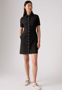Zwarte denim jurk met korte mouwen, knopen aan de voorkant, twee borstzakken en subtiele stikdetails. Model draagt vlakke witte schoenen.