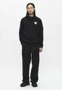 Schwarzer gestrickter Pullover mit rundem Halsausschnitt und weißem "W"-Emblem; kombiniert mit lockeren schwarzen Hosen und schwarzen Turnschuhen. Einfarbiger weißer Hintergrund.