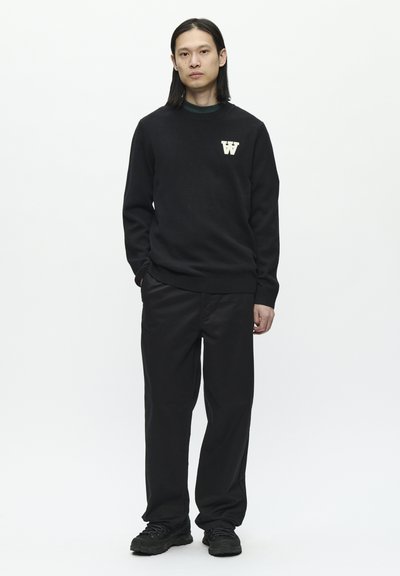 Suéter negro de punto con cuello redondo y emblema blanco con la "W"; combinado con pantalones negros holgados y zapatillas negras. Fondo blanco liso.