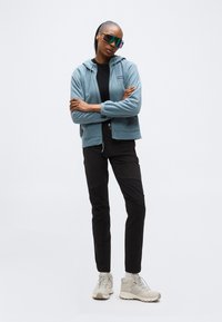 Γυναίκα με σταυρωμένα χέρια, φορώντας υπερμεγέθη φουτουριστικά γυαλιά ηλίου, μπλε φούτερ με φερμουάρ, μαύρο παντελόνι και μπεζ αθλητικά παπούτσια, στέκεται σε λευκό φόντο.