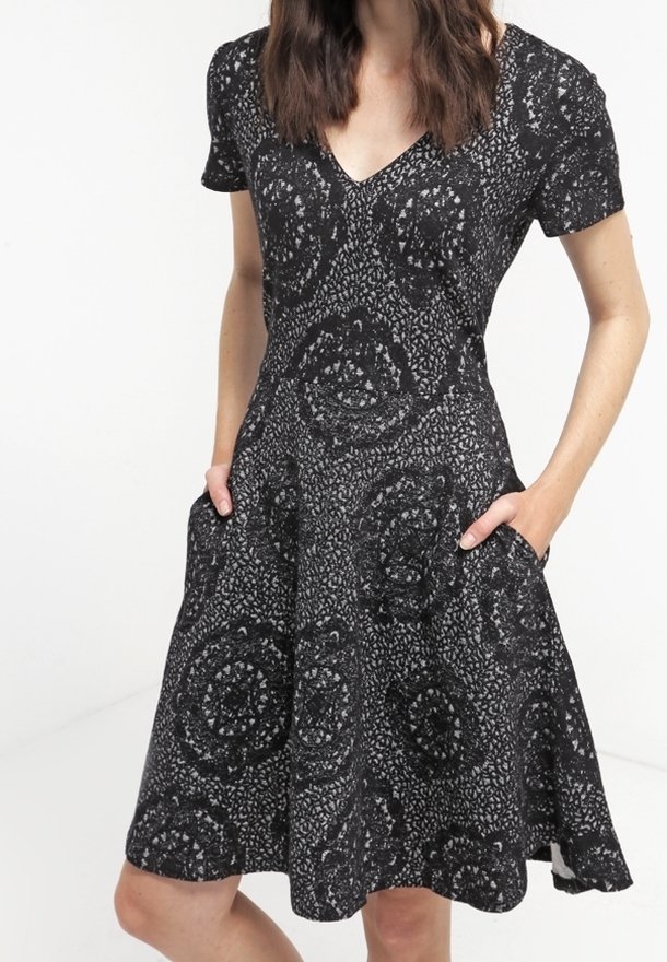 Schwarzes Kurzarmkleid mit V-Ausschnitt, featuring einem kreisförmigen Muster und Taschen. Der Stoff hat eine strukturierte Optik mit einem taillierten Oberteil und einem ausgestellten Rock.