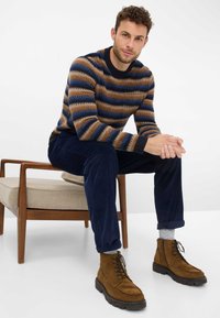 Gestreifter Pullover in Braun, Blau und Creme; kombiniert mit marineblauen Cordhosen und braunen Wildlederschuhen. Holzstuhl mit beigem Kissen im Hintergrund.