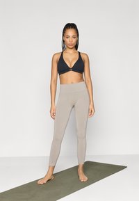 Μαύρο τριγωνικό αθλητικό σουτιέν και μπεζ ψηλόμεσα κολάν σε ένα μοντέλο, που στέκεται σε ένα πράσινο στρώμα γιόγκα σε μια απλή φόντο.