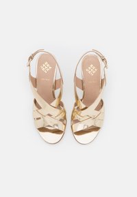 Sandales à talons hauts en cuir doré avec des brides croisées, design ouvert sur le devant et fermeture à boucle au niveau de la cheville. Texture lisse et brillante.