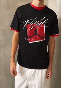 Černé bavlněné tričko s červenými akcenty, zdobené bílým textem "Flight" a černou siluetou skákajícího muže na červeném čtvercovém pozadí.