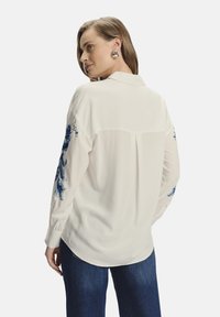 Blusa blanca con bordados florales azules en las mangas. Confeccionada en tejido ligero, con cuello y un corte relajado.