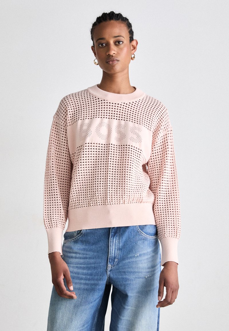 Pull en maille rose clair avec un design perforé, présentant un logo central "BOSS". Manches et ourlet ajustés, associé à un jean bleu décontracté.