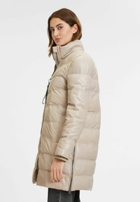 Giacca imbottita beige con colletto alto, chiusure a zip e tasche laterali. Presenta un design trapuntato e una texture liscia e lucida.