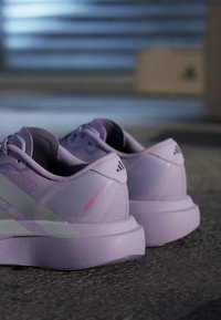 Para jasnofioletowych butów do biegania Adidas z białymi paskami i różowym tekstem "Adizero Pro SL" na betonowej podłodze w pomieszczeniu.