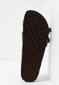 Sandal sål med et tekstureret, mørkebrunt gummimønster med cirkulære designs. Siden viser let brunt overmateriale med justerbare stropper.