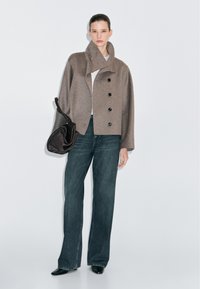 Giacca in misto lana marrone con grandi bottoni neri, dotata di un colletto alto. Abbinata a pantaloni in denim scuri a gamba larga e a una borsa marrone a forma curva.