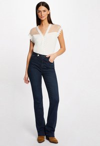 Blouse blanche avec des manches transparentes à pois et des bords festonnés, associée à un jean évasé bleu foncé et des bottines brunes.